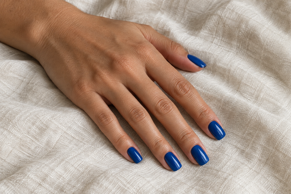 A hand wearing glossy royal blue press-on nails on soft ivory fabric.